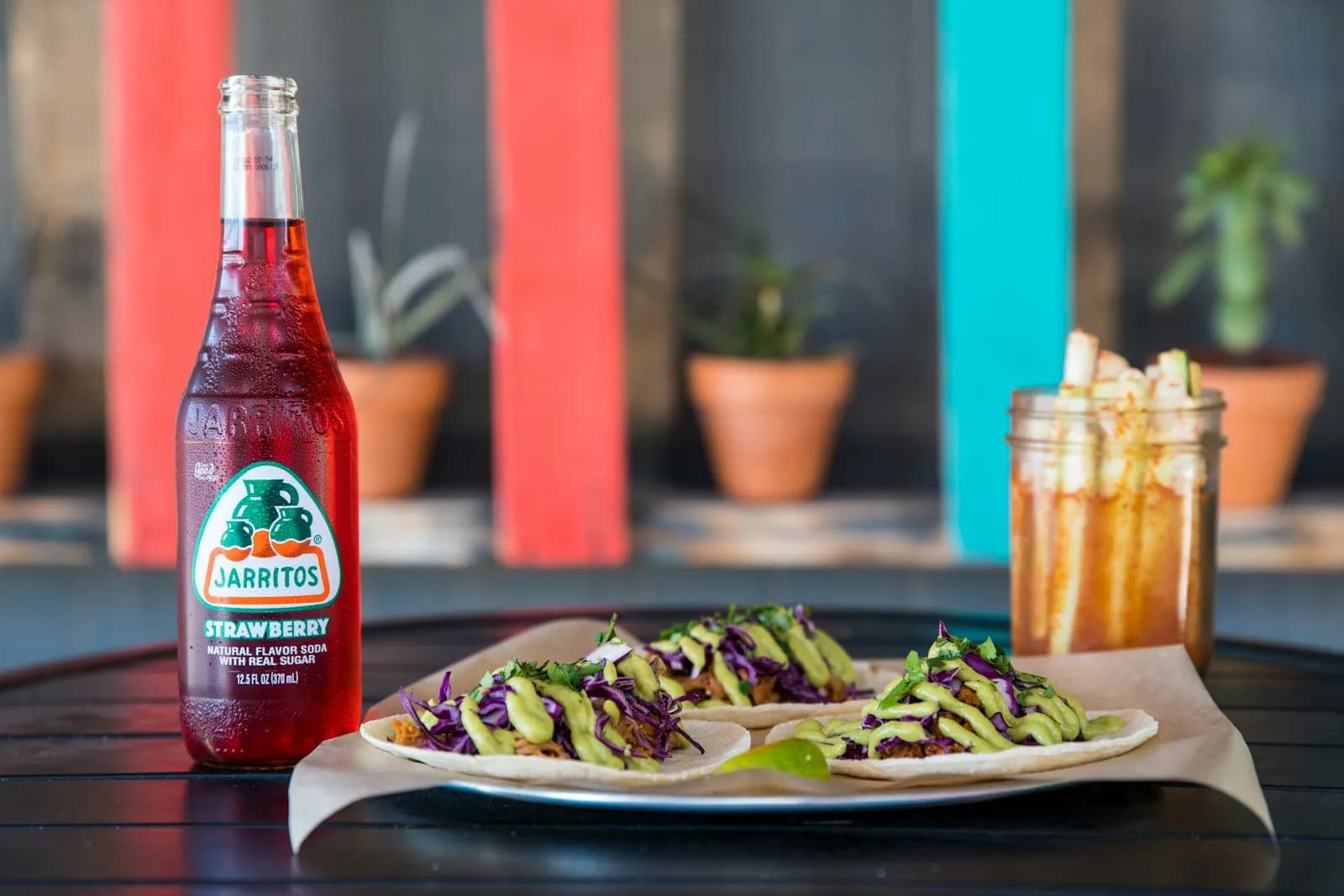 Tacos en un plato junto a una botella de refresco de fresa y un frasco con fruta en palitos