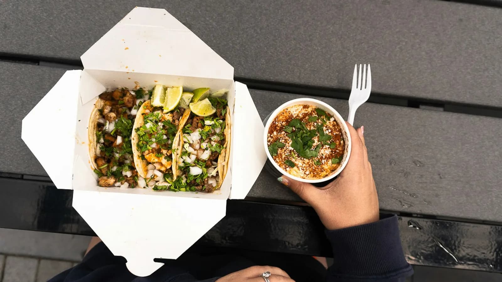 Vista desde arriba de una caja blanca con tres tacos de carne con cebolla y cilantro, un vaso de esquites en una mano y un tenedor de plástico sobre una banca oscura