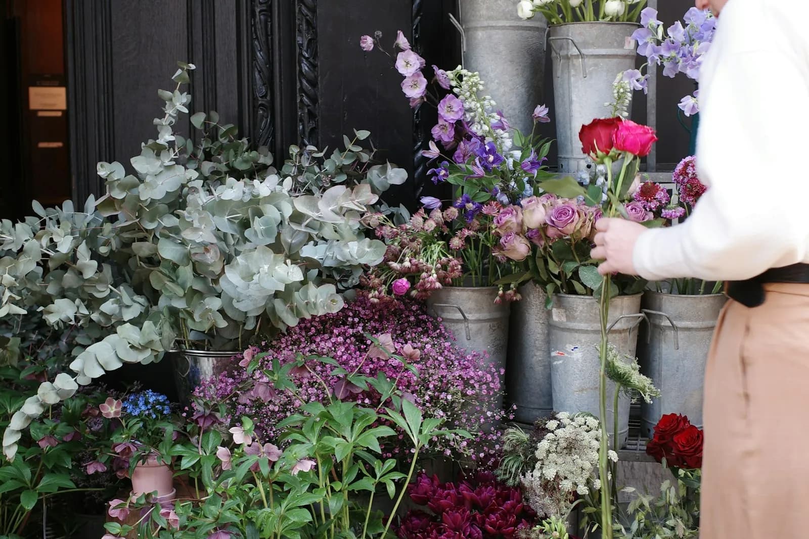 Una persona acomoda flores frente a cubetas con ramos de eucalipto y flores moradas