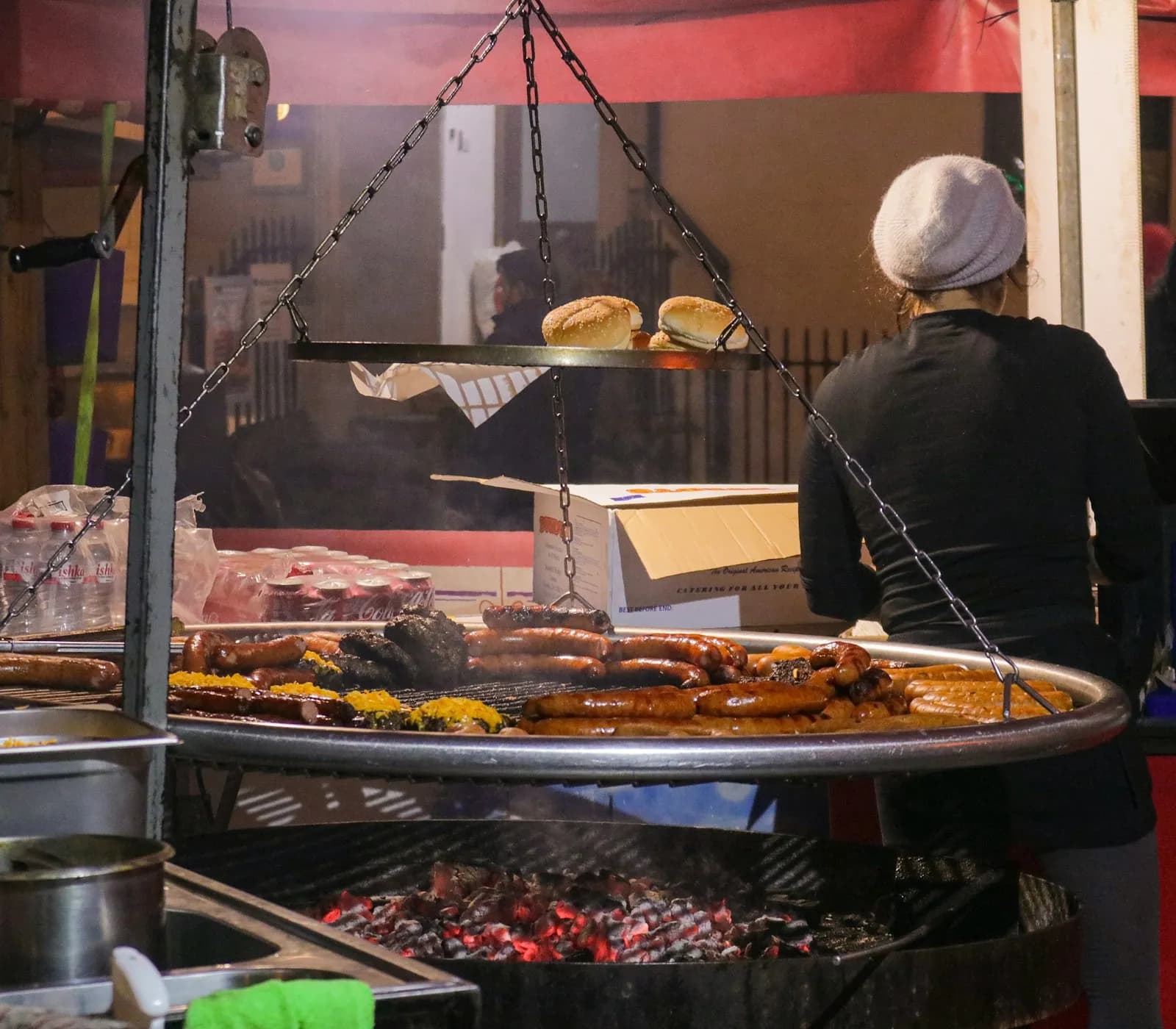 Persona con gorro frente a una parrilla redonda con salchichas y pan